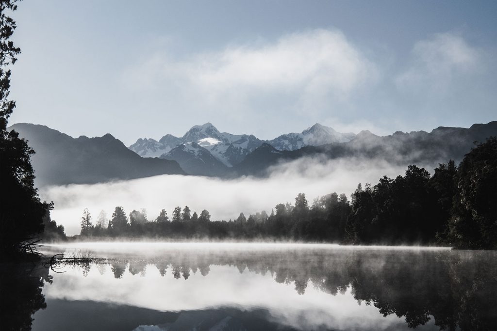 A beautiful sunrise at Lake Matheson in New Zealand with fog covering trees in the foreground and a beautiful mirror like reflection of mountains in the water.