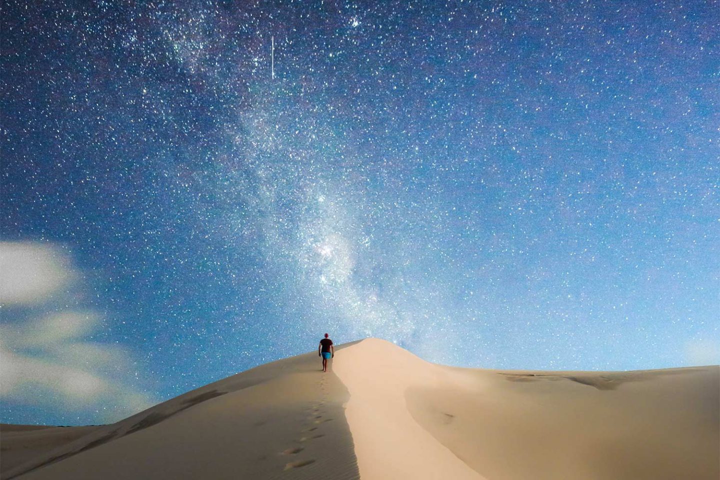 A man walks along sand dunes at night towards the Milky Way that lights up the night sky.
