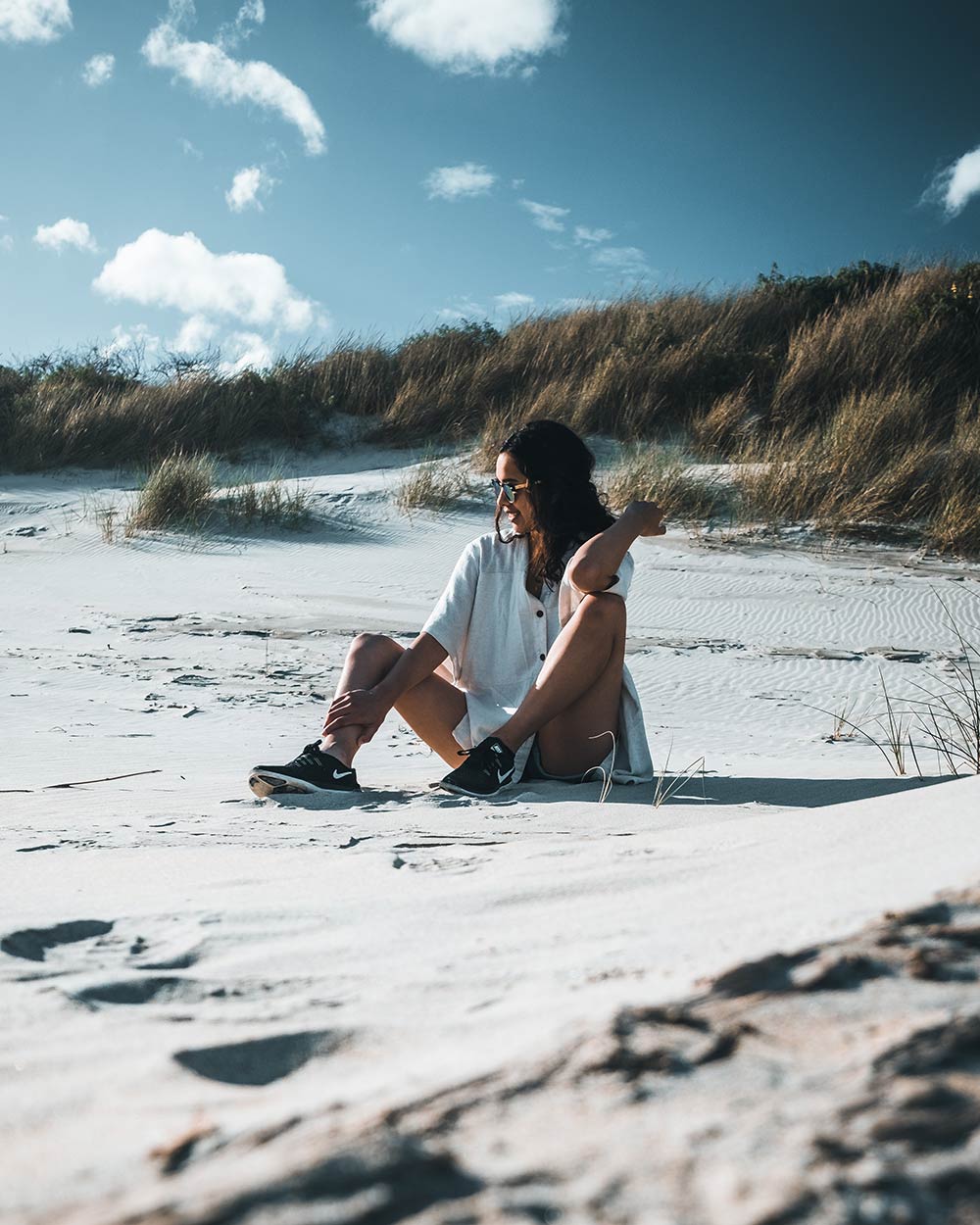 A woman sits on the sand of a beach with grassy hills and a beautiful blue sky behind her. This image has been edited using the Added Blues preset which is included as part of the moody Lightroom presets package.