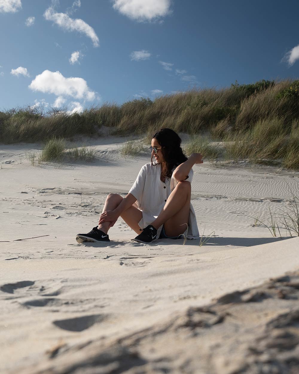 A woman sits on the sand of a beach with grassy hills and a beautiful blue sky behind her. This is the RAW unedited image without a preset applied.