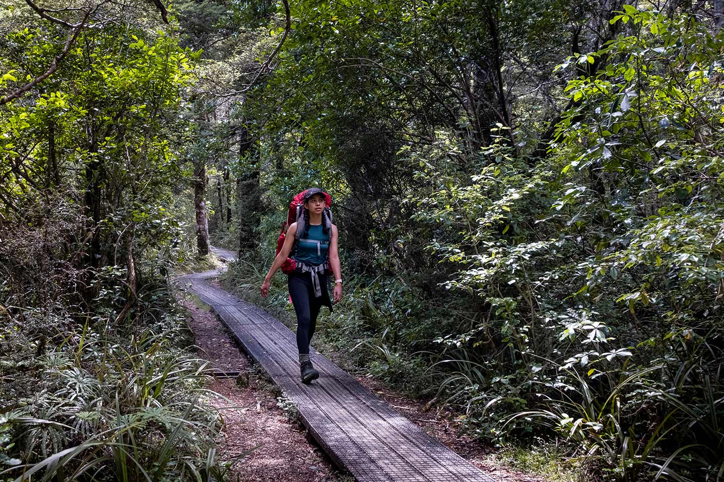A women walking along a wooden path with a red hiking pack on her back. She is surrounded by lots of lush green forest.